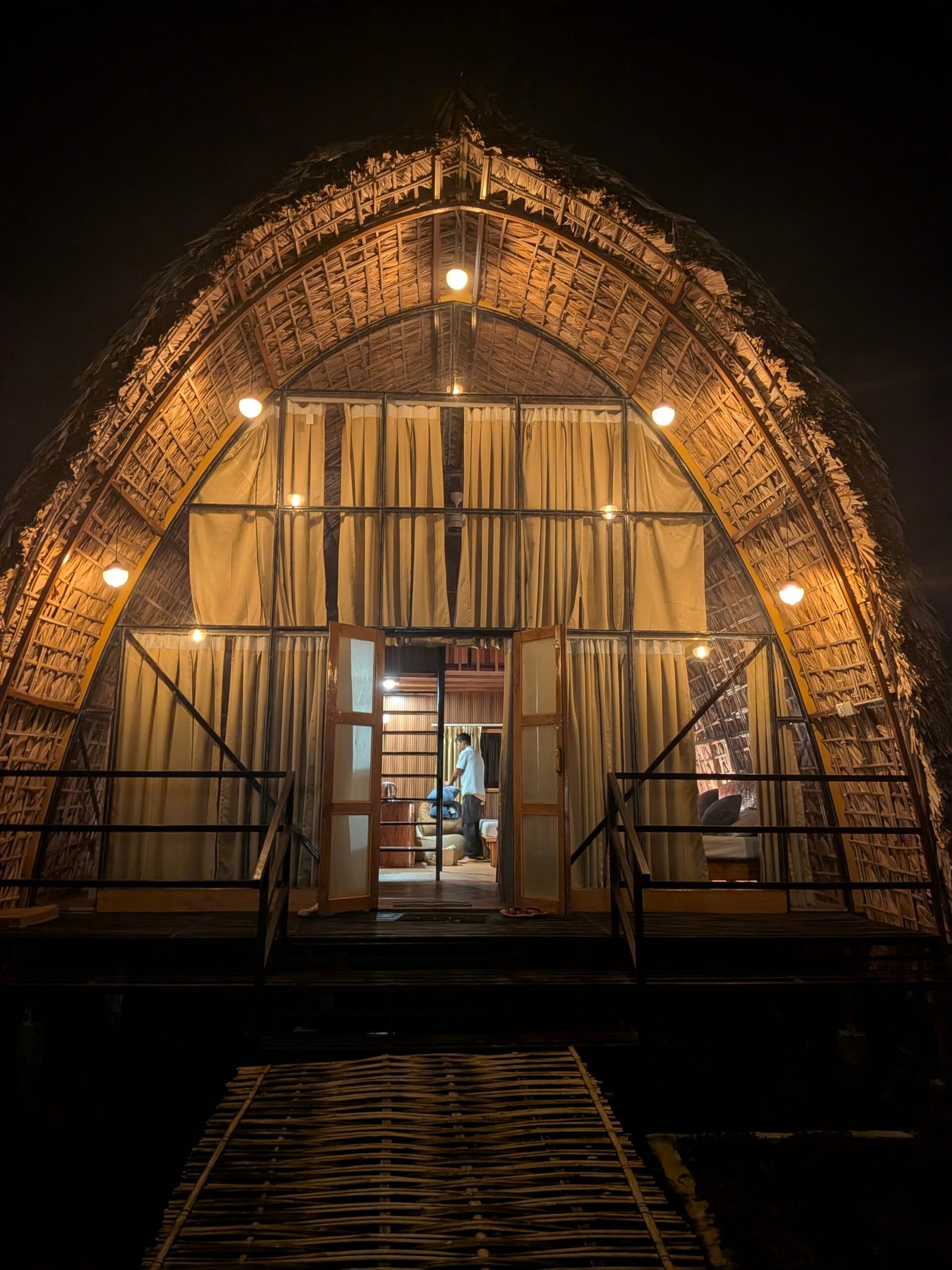 The Cocoon Cottage at Tushita Ban, lit and glowing at the end of a bamboo boardwalk through the grasslands at night